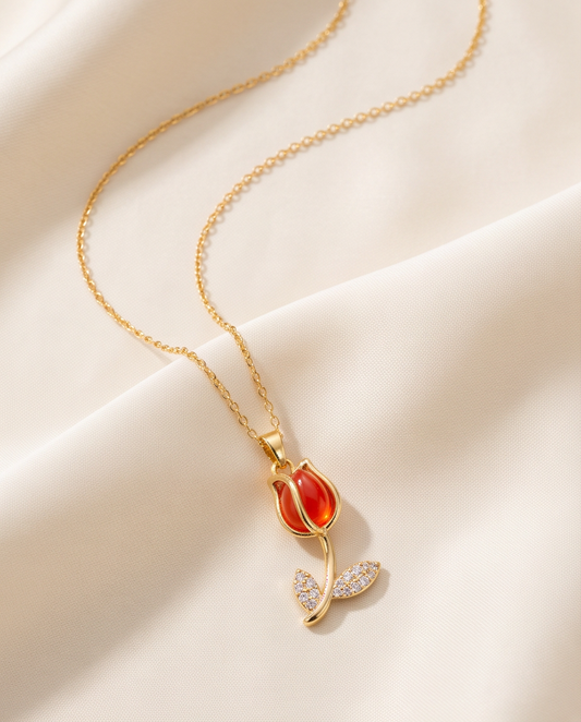 A gold chain necklace with a tulip pendant, featuring a glossy red-orange center and small clear crystal-encrusted leaves, displayed on draped cream satin fabric.
